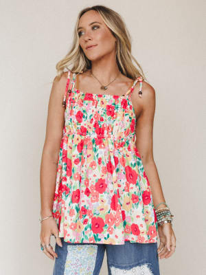 A breezy floral cami top with smocked bodice and tie straps, styled with patchwork jeans and layered bracelets against a soft neutral studio backdrop — the kind of top that makes a Tuesday feel like a garden party.