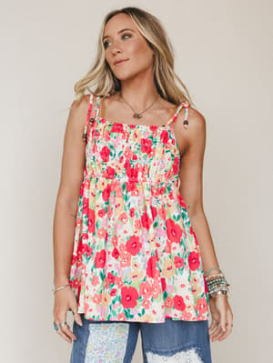 A breezy floral cami top with smocked bodice and tie straps, styled with patchwork jeans and layered bracelets against a soft neutral studio backdrop — the kind of top that makes a Tuesday feel like a garden party.