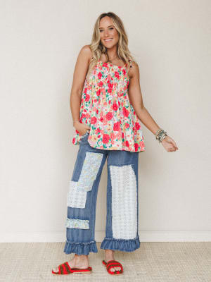 A smiling model in a breezy red floral smocked tank top paired with wide-leg patchwork denim jeans featuring eyelet and floral fabric inserts and ruffle hems — playful, warm, and full of personality against a soft neutral studio wall.