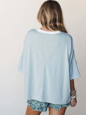 A back-view shot of an oversized striped tee in soft sky blue and white, paired with floral cutoff shorts and a stack of beaded bracelets — the kind of effortless warm-weather outfit that looks like it was thrown on in the best possible way.