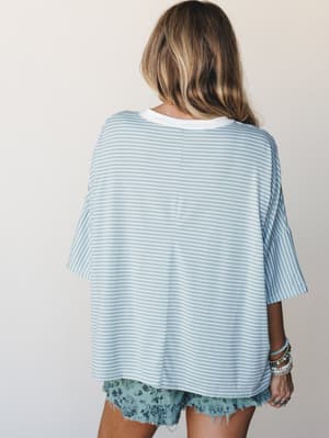 A back-view shot of an oversized striped tee in soft sky blue and white, paired with floral cutoff shorts and a stack of beaded bracelets — the kind of effortless warm-weather outfit that looks like it was thrown on in the best possible way.