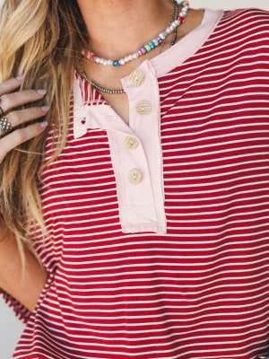 A close-up of a red and white striped henley top with a blush pink placket and natural wood buttons, styled with layered beaded necklaces and rings for a casual, collected everyday look.