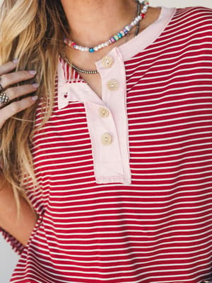 A close-up of a red and white striped henley top with a blush pink placket and natural wood buttons, styled with layered beaded necklaces and rings for a casual, collected everyday look.