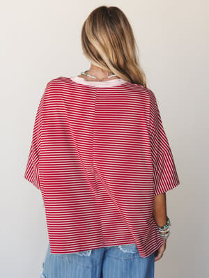 A back-view shot of an oversized striped top in marsala red and cream, styled casually over a layered pink tee with wide-leg denim — stacked bracelets and delicate necklaces round out the relaxed, lived-in feel.