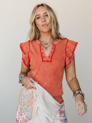 A sun-kissed ribbed tee in warm salmon with red contrast stitching and flutter sleeves, styled with patchwork floral pants and stacked boho jewelry — the kind of outfit that looks like it came together effortlessly but definitely didn't.