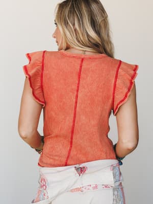 Shot from behind, a model in a salmon ribbed ruffle-sleeve tee with red contrast stitching is paired with white patchwork jeans — the kind of easy, expressive outfit that looks effortless but was absolutely thought about.