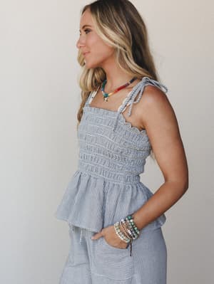 A smocked, tie-strap peplum top in a soft blue-and-white pinstripe pairs with matching wide-leg pants — styled with layered beaded bracelets and a rainbow beaded necklace for a playful, summery feel.