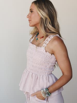 A side-profile shot of a model in a soft pink stripe smocked top with braided straps, layered with colorful beaded jewelry — the kind of outfit that makes a Saturday feel intentional.