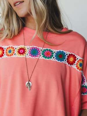 A coral relaxed tee with a bold granny square crochet panel across the chest — the kind of top that makes a Tuesday feel a little more colorful, styled with a long feather pendant necklace.