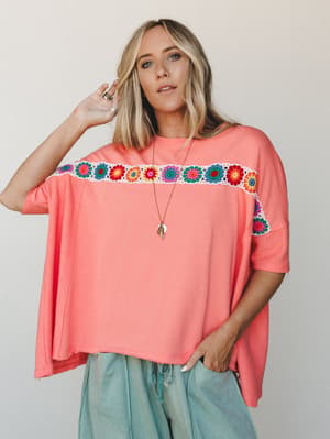 An oversized coral tee with a bold granny square crochet band across the chest, styled with sage drawstring pants and a long pendant necklace — the kind of top that looks like it tried zero percent and landed at one hundred.