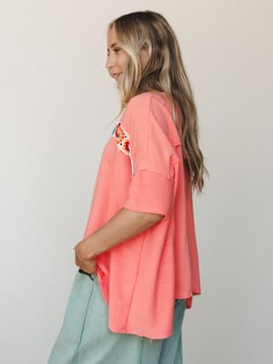 A relaxed coral oversized tee shot from a side-back angle, showing off a colorful crochet granny square patch at the shoulder — paired with sage wide-leg jeans for an effortlessly lived-in look.