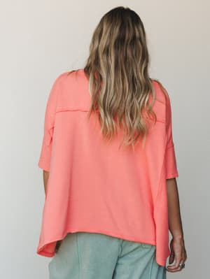 A back-view shot of a relaxed, boxy coral tee with raw-edge seam detailing, paired with washed sage jeans — the kind of effortless combination that feels intentional without trying.