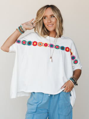 An oversized off-white tee with a bold multicolor crochet floral trim across the chest and sleeve, styled with light blue cargo pants and layered boho jewelry — the kind of top that does all the talking so you don't have to.
