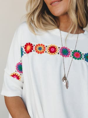 A relaxed white tee with a vibrant granny square crochet band across the chest and sleeves — the kind of top that does the talking for you, styled with a layered feather charm necklace for an effortlessly personal touch.