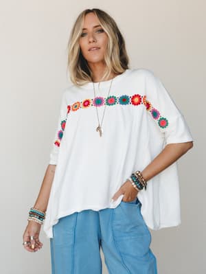 An oversized off-white tee with a colorful crochet floral trim across the chest and sleeves, styled with wide-leg light wash cargo pants and layered boho jewelry — the kind of outfit that looks effortless but totally intentional.
