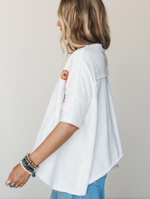 A side-back view of a relaxed white fleece tee with floral embroidery on the sleeve, styled with stacked boho bracelets and light blue jeans — the kind of top that looks like it was made for slow mornings and spontaneous plans.