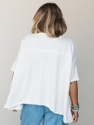 A back-view shot of an oversized white tee with exposed seam detailing and a raw hem, paired with light blue jeans and a stack of beaded bracelets — effortless and lived-in in the best way.