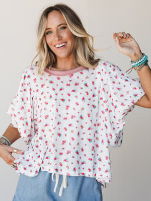 A relaxed, oversized floral top in ivory with a ditsy rose print and contrasting pink ribbed neckline — styled with light blue bottoms and stacked turquoise bracelets for that effortlessly happy, warm-weather kind of energy.