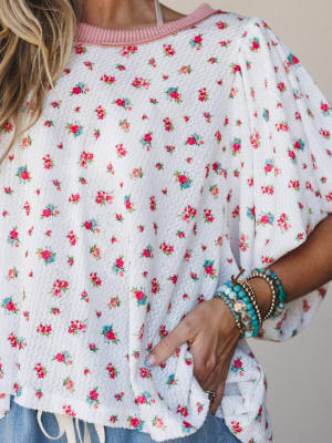 A close-up of a floral printed waffle-knit top in ivory with a pink ribbed neckline and relaxed ruffle sleeves, styled with stacked beaded bracelets and a peek of denim — effortlessly springy and a little nostalgic.