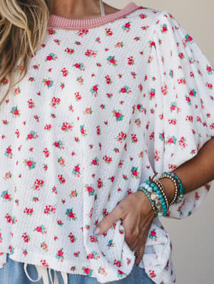 A close-up of a floral printed waffle-knit top in ivory with a pink ribbed neckline and relaxed ruffle sleeves, styled with stacked beaded bracelets and a peek of denim — effortlessly springy and a little nostalgic.