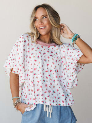 A flowy ivory floral top with ruffle sleeves and a drawstring hem, styled with blue wide-leg pants and stacked boho bracelets — the kind of top that makes a Tuesday feel intentional.
