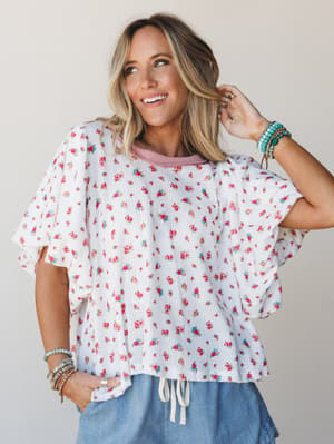 A flowy ivory floral top with ruffle sleeves and a drawstring hem, styled with blue wide-leg pants and stacked boho bracelets — the kind of top that makes a Tuesday feel intentional.