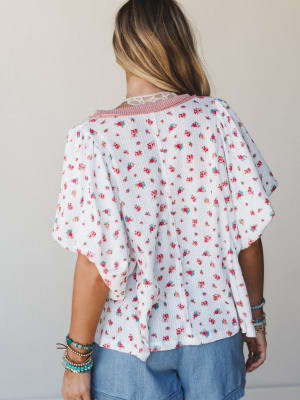 A back-view shot of a flowy ditsy floral top in ivory with flutter sleeves and a pretty lace and striped collar detail, styled with chambray shorts and stacked boho bracelets — the kind of top that makes a Tuesday feel intentional.