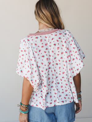 A back-view shot of a flowy ditsy floral top in ivory with flutter sleeves and a pretty lace and striped collar detail, styled with chambray shorts and stacked boho bracelets — the kind of top that makes a Tuesday feel intentional.
