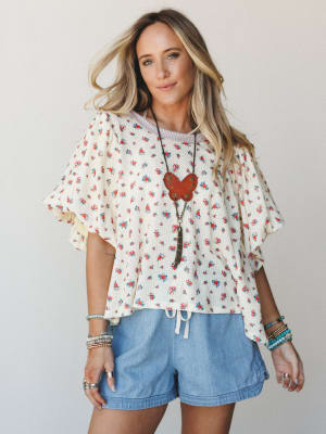 A breezy floral ruffle-sleeve top in cream with ditsy rose print is styled with light chambray shorts, layered boho jewelry, and a statement leather pendant — effortless and a little nostalgic, like a good summer afternoon.