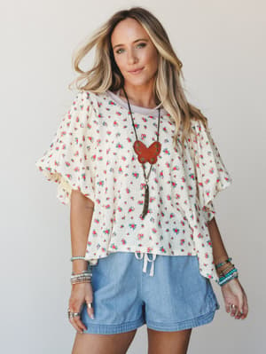 A breezy floral ruffle-sleeve top in cream with ditsy rose print is styled with light chambray shorts, layered boho jewelry, and a statement leather pendant — effortless and a little nostalgic, like a good summer afternoon.