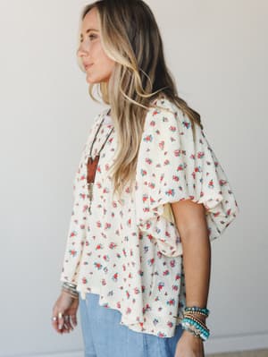 A breezy floral kimono-style top with dramatic ruffle sleeves worn over a graphic tee and denim, styled with turquoise beaded bracelets and a boho pendant necklace — the kind of outfit that feels effortless but clearly thought about.