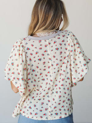A back-facing shot of a cream floral top with fluttery ruffle sleeves and a delicate lace neckline detail — the kind of piece that looks just as good from behind as it does from the front.