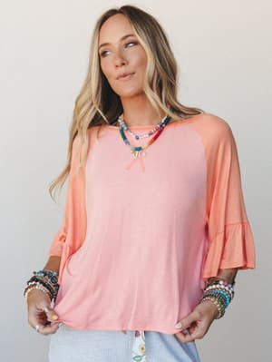 A relaxed two-tone top in blush pink and peach tangerine with flutter bell sleeves, styled with stacked beaded bracelets and layered colorful necklaces — the kind of effortless, happy outfit that practically plans the rest of your day.