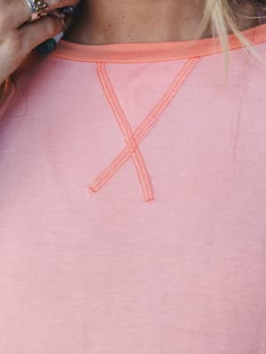 A close-up detail shot of a soft pink top with a coral contrast neckband and distinctive cross-stitch seam detail at the chest — the kind of thoughtful construction detail that makes a simple top feel anything but.