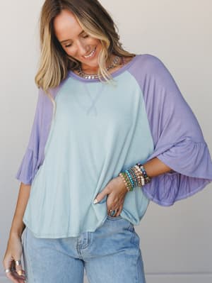 A relaxed color-block top in soft sky blue and lavender with fluttery bell sleeves, styled with light wash jeans and a stack of boho bracelets — the kind of top that makes a Tuesday feel a little more like itself.