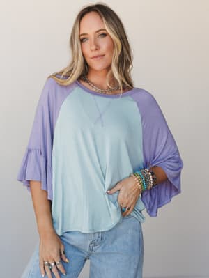 A relaxed colorblock raglan top in soft sky blue and lavender with flutter bell sleeves, styled with light-wash jeans and layered boho jewelry for an effortlessly personal, everyday kind of feel.