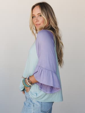 A side-profile shot of a colorblock top in soft sage and lavender, styled with light-wash jeans and a stack of beaded bracelets — the kind of easy, expressive outfit that looks like you tried just enough.
