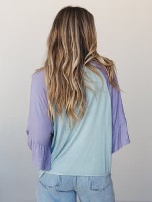 Shot from behind, a model in a colorblock top pairs soft sage and lavender in a relaxed silhouette — the ruffle bell sleeves add just enough movement to make it feel intentional rather than casual.