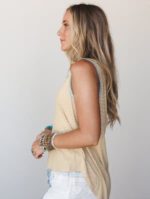A side-profile shot of a model in a mustard and white striped tank top with a soft blue trim neckline, styled with white patterned shorts and a stack of colorful beaded bracelets — relaxed and summery with an easy boho feel.
