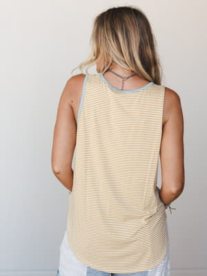 A back-view shot of a relaxed striped tank in warm mustard and white, with a soft blue contrast trim at the neckline and armholes — easy, sun-worn, and made for wherever summer takes you.