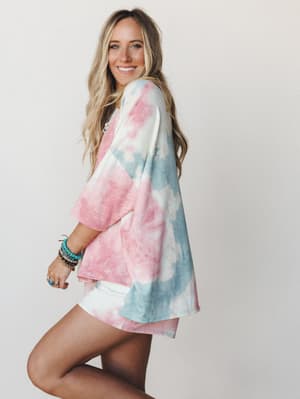A smiling model poses in a side profile wearing an oversized tie-dye top in cotton candy pink and dusty blue over white shorts, styled with stacked turquoise bracelets and silver rings — the kind of effortless, happy outfit that makes a regular day f