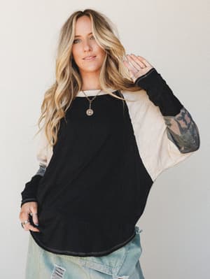 A relaxed colorblock long-sleeve top with a black body, oatmeal sleeves, and camo cuffs — worn with distressed light-wash jeans and layered gold necklaces for that effortless, lived-in weekend vibe.