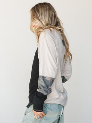 A smiling model shot from behind wearing a color-block long-sleeve top with a cream body, black sleeve, and camo-print sleeve panel, paired with distressed light-wash jeans and silver rings — easy, lived-in style with a little edge.