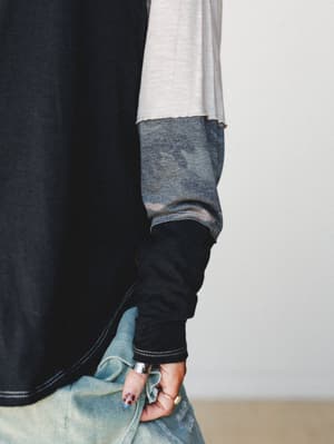 A close-up detail shot showing intentional layering — a black oversized top over a cream thermal and camo sleeve underneath, styled with light wash distressed denim and gold rings, capturing that effortless lived-in look.
