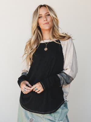 A relaxed colorblock long-sleeve top with a black body, cream sleeves, and camo cuffs worn with distressed light-wash jeans and layered boho jewelry — effortless and lived-in with just enough personality.