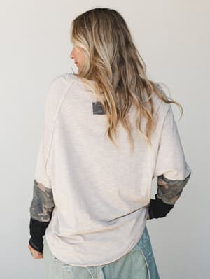 Shot from behind, a model in a relaxed oatmeal long-sleeve top with camo-print contrast sleeves and black ribbed cuffs pairs it with distressed light-wash jeans — the kind of effortless layered look that feels collected without trying.