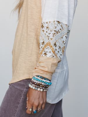 A close-up detail shot highlighting the patchwork-print sleeve of a colorblock long-sleeve top styled with a stack of beaded bracelets and silver rings — earthy, collected, and very much the kind of arm you notice across the room.