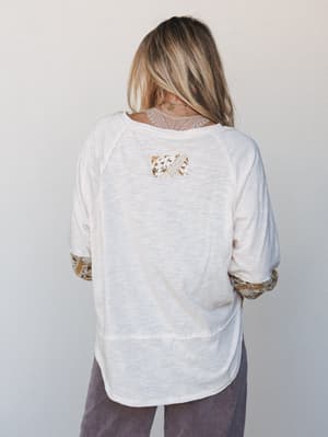 Shot from the back, a relaxed cream slub-knit top with mixed-fabric detailing — patchwork panel at center back, lace inset at the neckline, and printed cuffs — layered over dusty mauve wide-leg pants, giving off that effortless 'threw this on and som