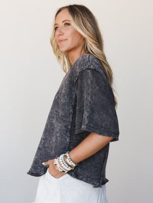 A side-profile shot of a model in a boxy, acid-washed charcoal crop top with raw-edge hem details, styled casually with white pants and a stack of beaded boho bracelets — the kind of effortless piece that looks intentional without trying.