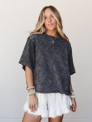 An oversized acid-wash crop tee in charcoal pairs effortlessly with frayed white drawstring shorts, layered bracelets, and delicate gold jewelry — the kind of outfit that looks like you tried just enough.